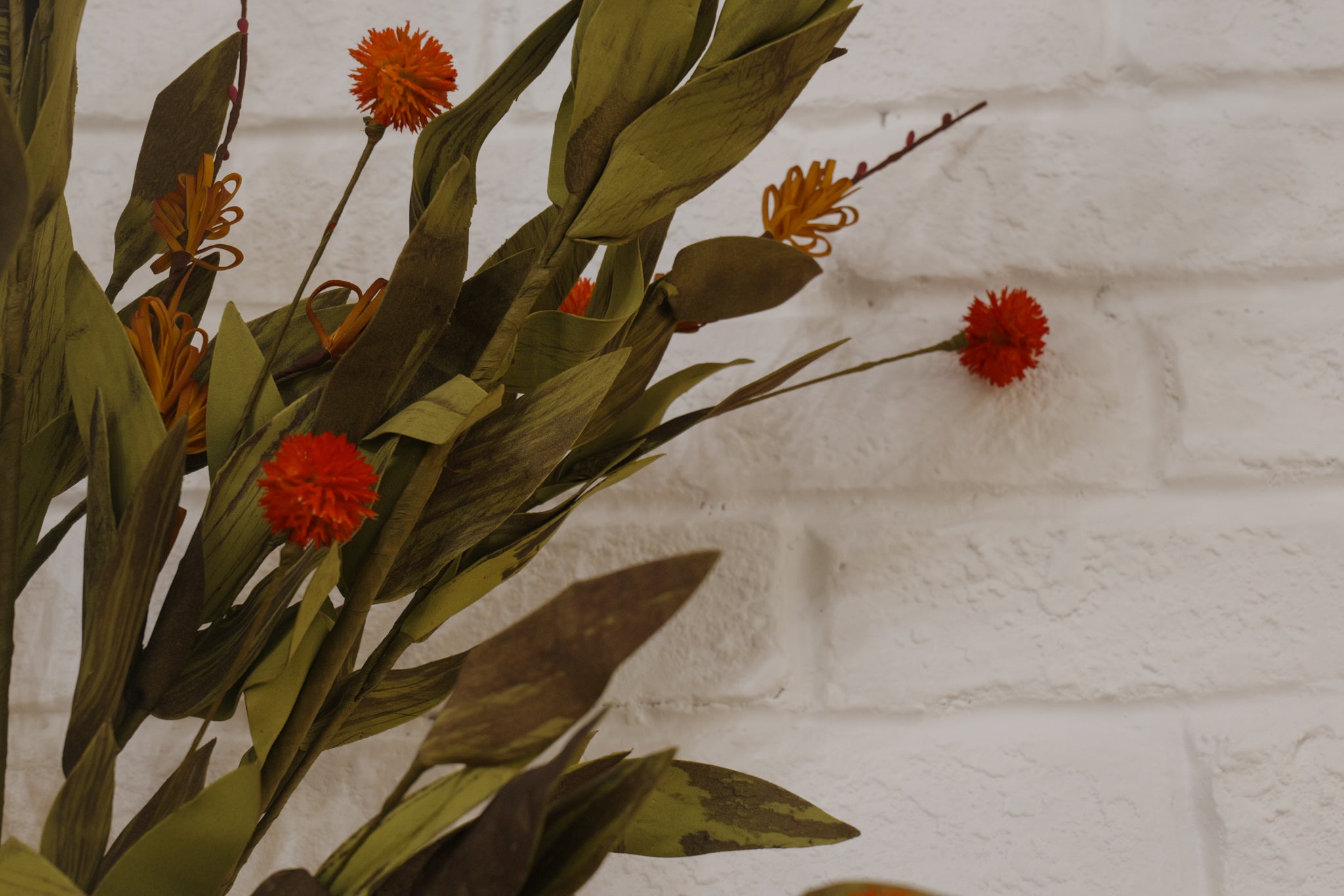 Artificial plants with red flowers and green leaves against a white brick wall.