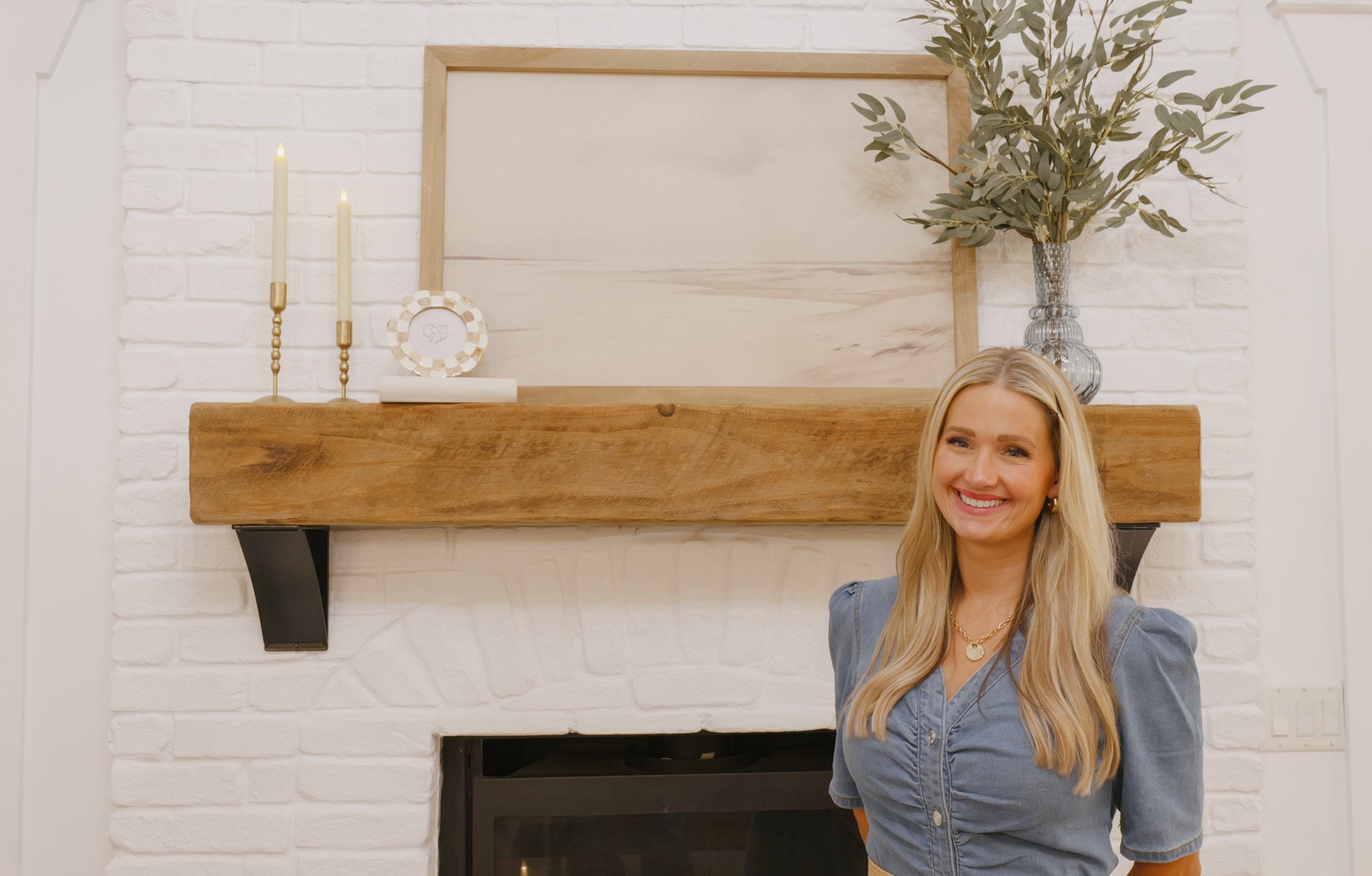 Woman standing in front of a fireplace with a wooden mantel and decorative items.