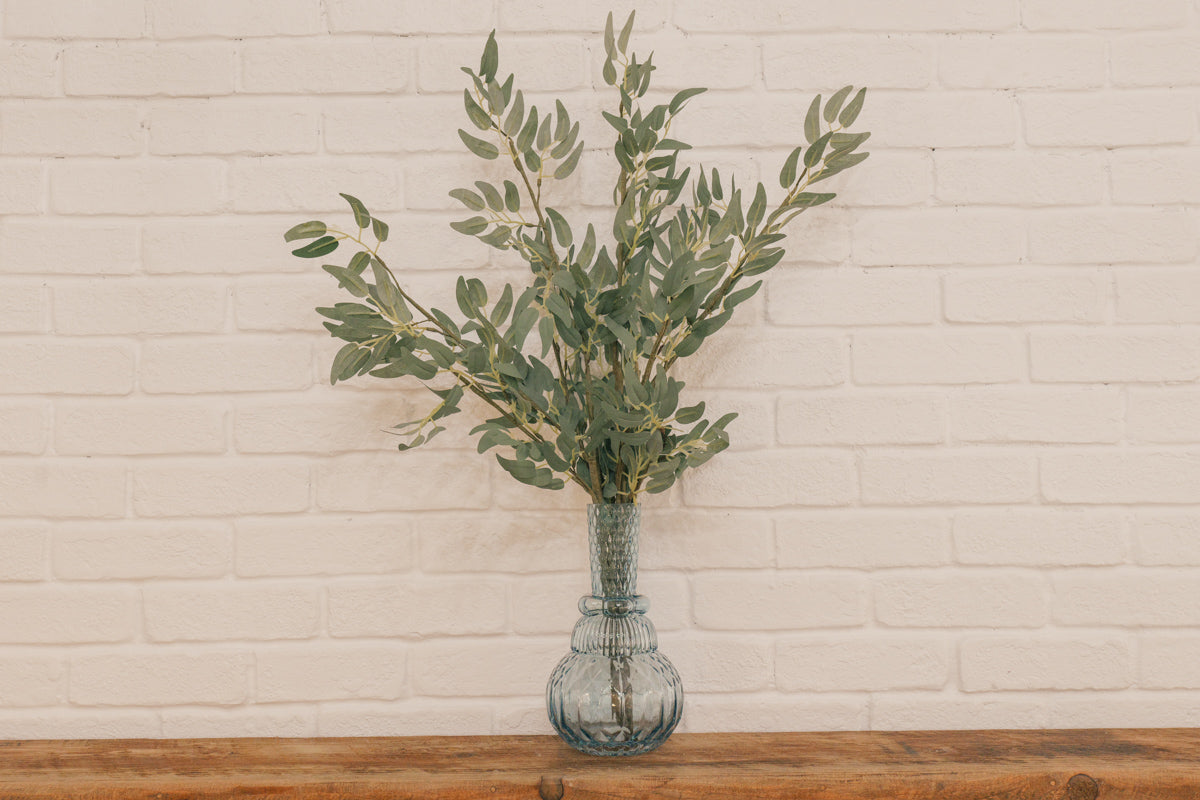 Vase with greenery on a wooden surface against a white brick wall