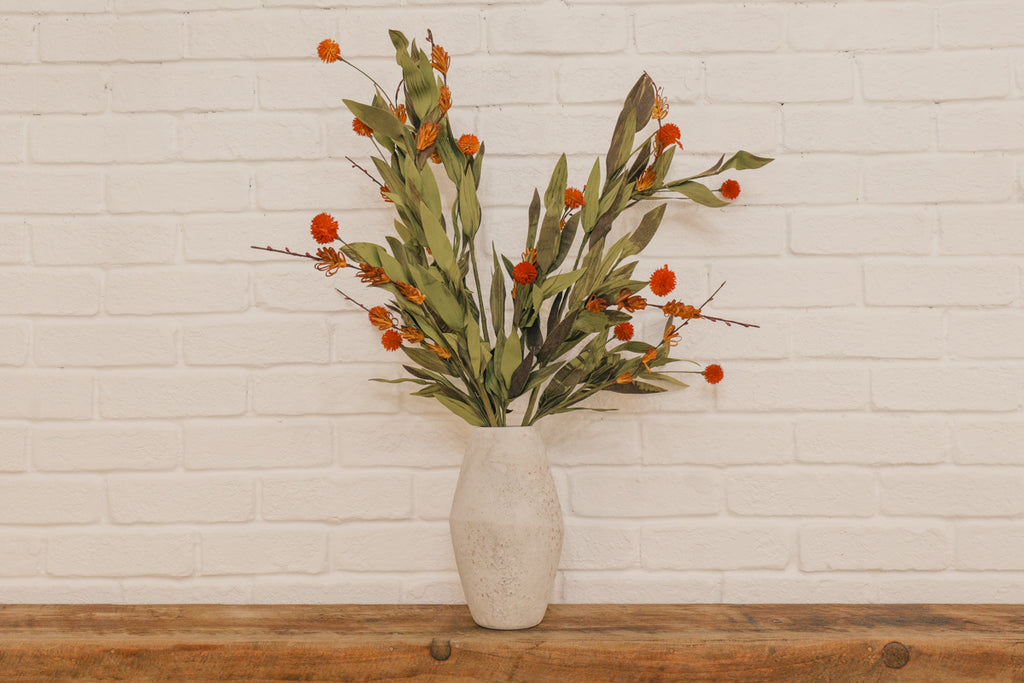 Vase with greenery and orange berries on a wooden surface against a white brick wall.