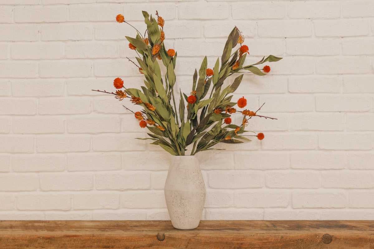 Vase with greenery and orange berries on a wooden surface against a white brick wall.